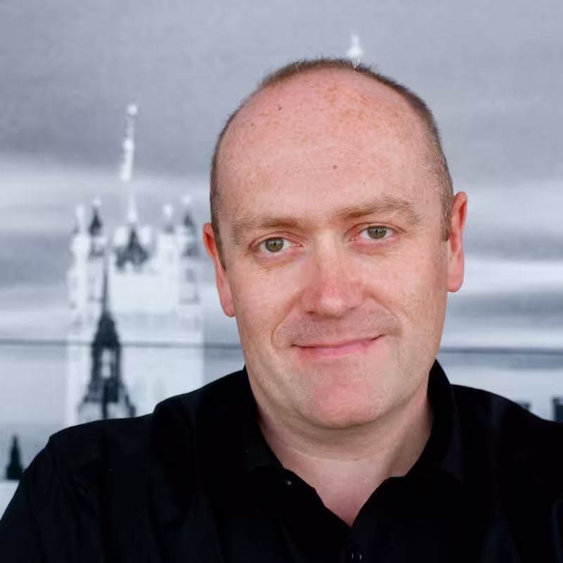 Cennydd: a black and white cut-out photo of a smiling man with a shaved head, wearing a dark shirt
