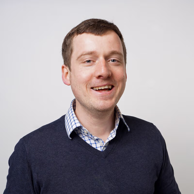 Hidde: a man with short brown hair smiling broadly at the camera, wearing a navy jumper over a blue and white checked shirt against a light grey background
