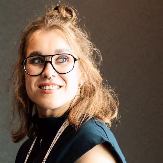 Lena: a woman with shoulder length hair, a nose ring and bold full rim 70s style glasses whilst looking off to the side