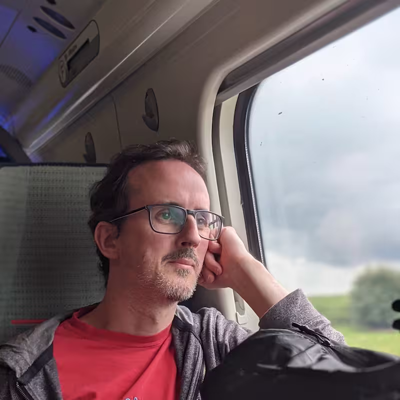 Matthew: a man with short dark hair, glasses and a greying beard, resting his head on his hand and gazing out of a train window at green fields, wearing a red t-shirt under a grey hoodie