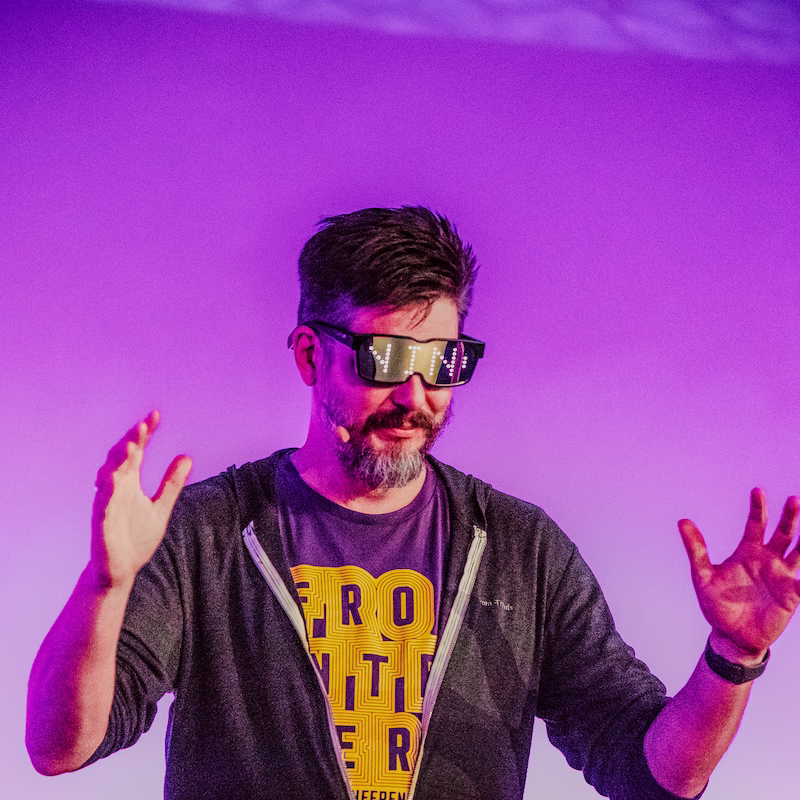 Under vibrant purple stage lighting, a bearded man wearing LED-illuminated matrix glasses that reads "NIN" and a dark zip-up hoodie over a graphic t-shirt presents animatedly with both hands raised.